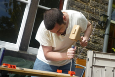 Dallas happily crafting a wooden piece outdoors, focused and enjoying his work at the bench.
